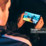 Man holds mobile in his hands and play a mobile action game. It features a crosshair and a machine gun. He sits at home at night playing the game.