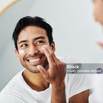 Man applying face skincare cream while grooming looking at the mirror in the bathroom in his morning routine at home. Close up portrait of handsome guy using facial lotion for clean and clear skin