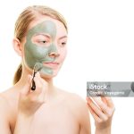 Beauty skin care cosmetics and health concept. Young woman holding brush and bowl with facial clay mask applying mud to her face isolated