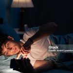 Portrait of young sleepy man lying in bed using smartphone at late night, no sleep. Insomnia, sleep disorder concept. Dependency on a cellphone. Loneliness crisis. Copy space